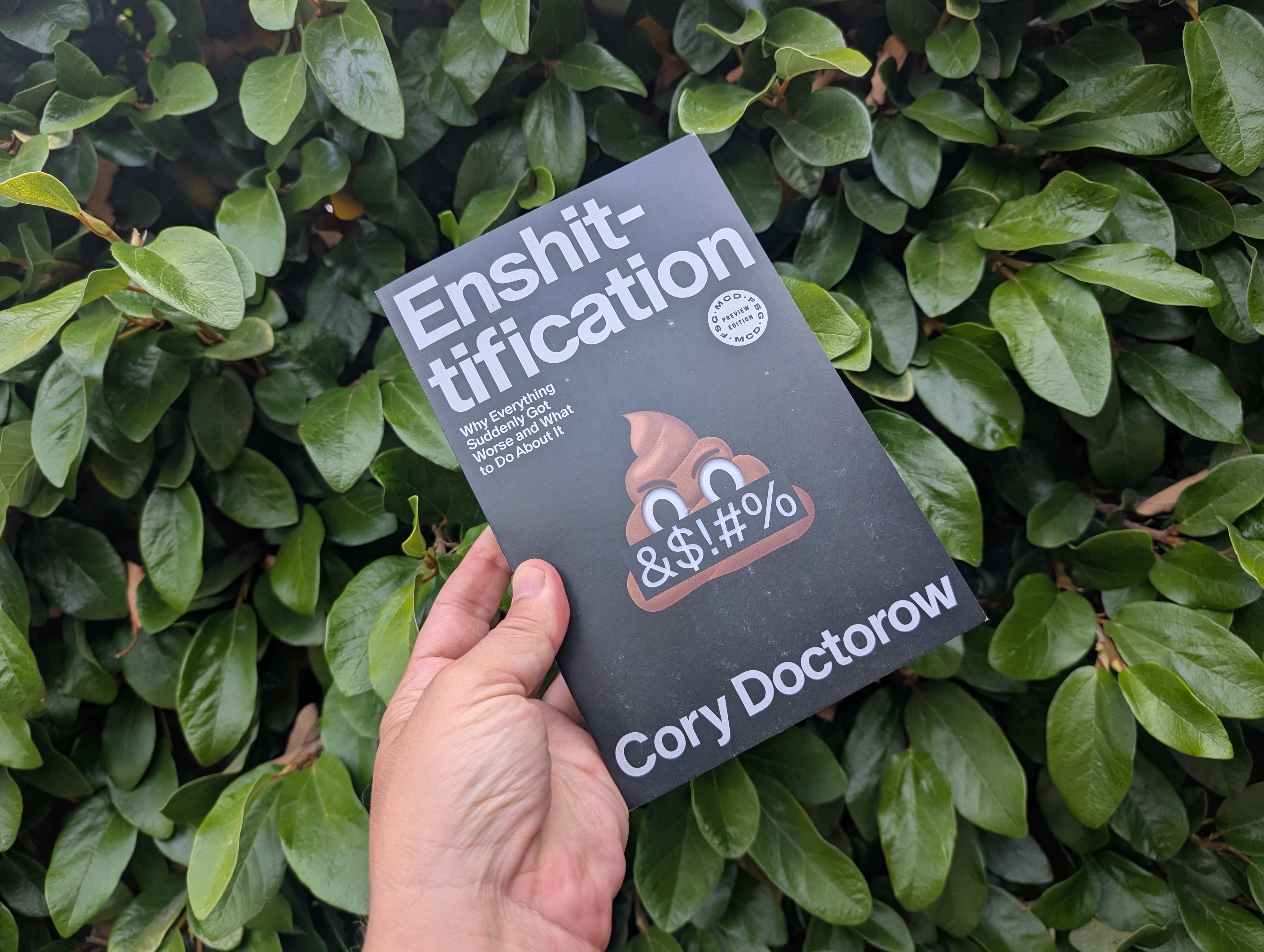 Hand holding the paperback of the book in front of a hedge